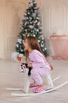 A beautiful little girl in pink pajamas rejoices in a wooden rocking horse, a gift from Santa for Christmas.