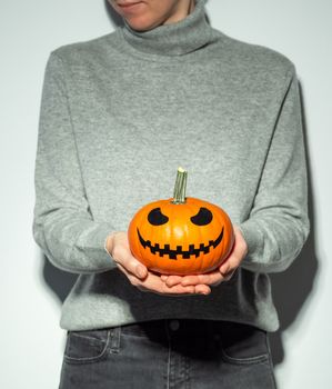 Crop unrecognizable woman holding halloween scary face bright orange pumpkin. Young woman in gray cashmere sweater and black jeans with jack-o-lantern pumpkin in hands. Snapshot style.. Copy space