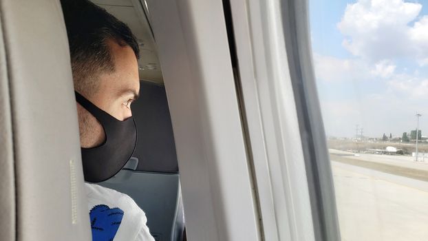 Spy photo through the seat, a masked guy on the plane looks out the window. Air travel during the coronavirus pandemic.