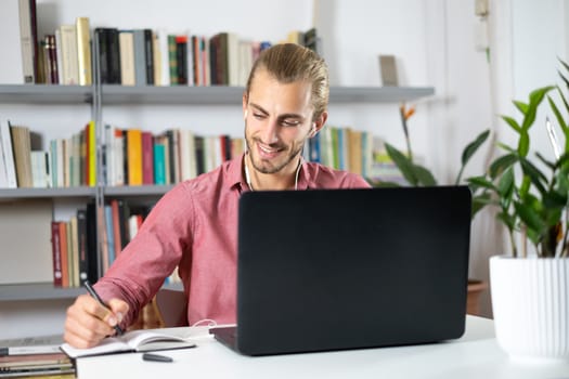 Attractive young man sitting at the table at home working with a laptop with happy face and smiling with a smile and writing on the book