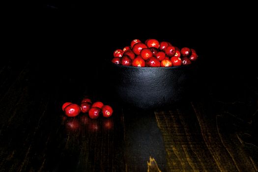 A bunch of cranberrries in a black, cast iron bowl, with black background and brown base. Shot in low key style.