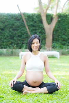 Beautiful pregnant asian young woman relax in the park, girl with belly sitting with crossed leg on grass and yoga exercise for wellness, copy space.