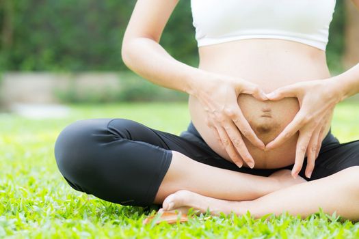 Happy pregnant beautiful young woman with gesture hand heart shape in the park, closeup woman with hand touch belly with relax, mother love baby, family concept.