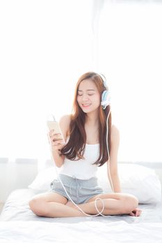 Beautiful asian young woman enjoy listen music with headphone and holding smart mobile phone while sitting in bedroom, relax girl with earphone, leisure and technology concept.