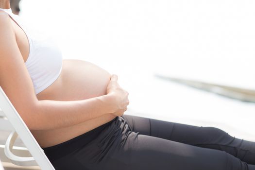 closeup beautiful pregnant asian young woman relax in the park, girl with belly sitting at swimming pool for wellness, copy space.