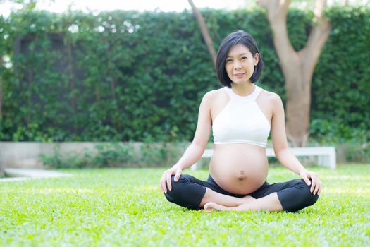 Beautiful pregnant asian young woman relax in the park, girl with belly sitting with crossed leg on grass and yoga exercise for wellness, copy space.