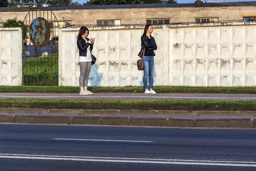 Belarus, Minsk - September 09, 2017: two girls are standing on the sidewalk near the fence. One girl looks into the distance, the second girl looks into the smartphone