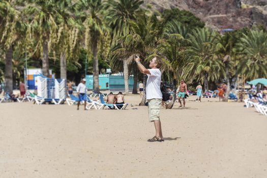 Spain, Tenerife - September 12, 2016: Man standing on the beach and taking pictures on smartphone