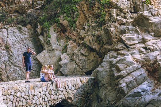 Spain, Lloret de Mar - September 22, 2017: Two girls and a guy on a stone bridge near the mountain, the girls are sitting, the guy is standing.