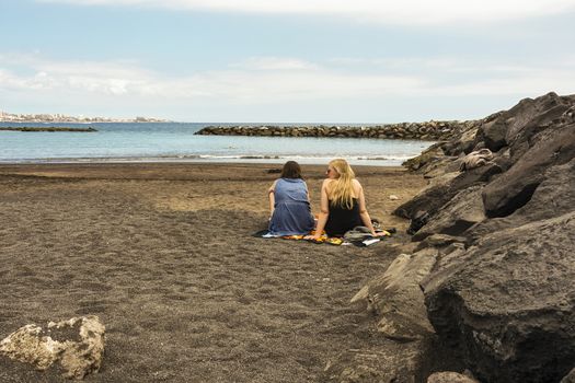 Spain, Tenerife - May 10, 2018: Two young women sit on the sand on the ocean and talk