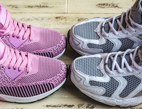 On the wooden surface is a pair of pink women's sneakers and a pair of white men's sneakers.
