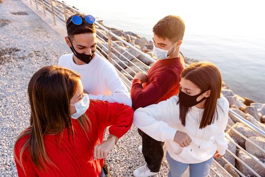 Top view of a group of young friends wearing protective face masks greeting bumping elbows outdoor Near the sea rocks in a vacation place in Sardinia - New normal contacts with social distancing