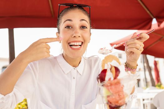 Beautiful smiling happy young caucasian woman doing yummy gesture with a finger while eating a big compound ice cream with fruit - New alimentary bad human habits of gluttony people