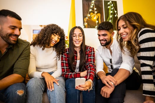 Group of multi ethnic young friends at home sitting on the sofa having fun looking at the smartphone - Caucasian woman with glasses and plaid shirt using social network technology to sharing