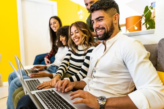 Group of multi ethnic young friends at home sitting on the sofa using three aluminum laptop - Side view of lined up millennials working and having fun with new technology - Focus on the first man