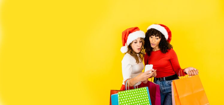Two attractive women with Santa hat looking to a smartphone excited and surprised - Big yellow banner with female people doing shopping online holding colored shopping bags on yellow big copy space