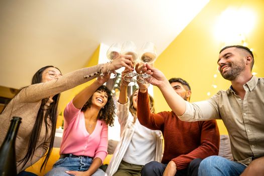 Bottom view of a multi ethnic young friends group at home sitting on the sofa toasting with white wine glasses - Multiracial happy people having fun drinking champagne for an event celebration