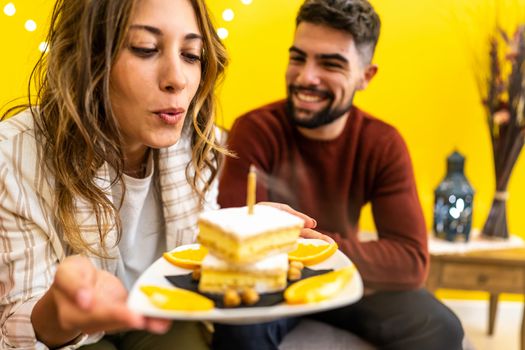 Young cute blonde woman blows out the candle of her birthday cake with her smiling boyfriend sitting on the sofa - Couple event celebration at home - Focus on the tuft of hair in the center