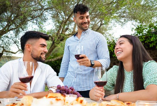 Three attractive young happy people celebrating outdoor holding red wine glasses - Stylish millennial friends having fun in the garden drinking alcohol at a table laden with snacks and grapes