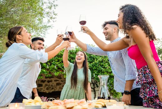 Multi ethnic happy young friends group toasting in the garden at sunset with red wine glasses - Millennials having fun together in countryside celebrating the grape harvest in Tuscany