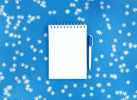 White blank sheet of notebook and a pen on blue background with scattered confetti snowflakes. Holiday education concept. Stock photo.