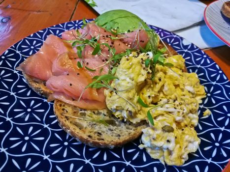 Smoked salmon and healthy scramble eggs on brown hard toast on blue plate in a cafe