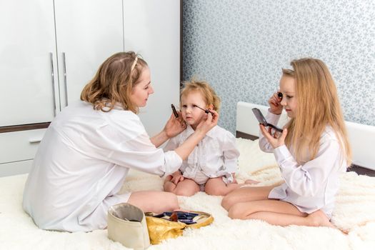 Portrait of mom and her daughters doing makeup to each other in the bedroom on the bed.