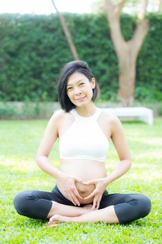 Happy pregnant beautiful young asian woman with gesture hand heart shape in the park, woman with hand touch belly with relax, mother love baby, family concept.