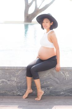 Beautiful pregnant asian young woman relax in the park, girl with belly sitting at swimming pool for wellness, copy space.