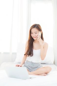 Beautiful young asian woman using laptop and talking smart mobile phone for leisure on bedroom, girl working online with notebook freelance, business concept.