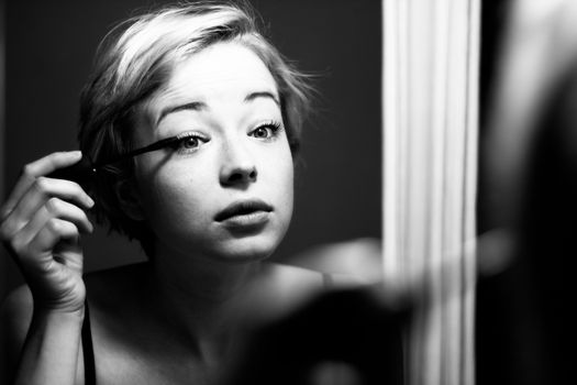 Woman getting ready for work doing morning makeup routine applying mascara in bathroom mirror at home. Beautiful caucasian girl applying eye make-up. Black and white image.