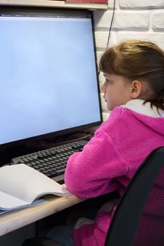 A girl studies at a computer with a large monitor, view from the back