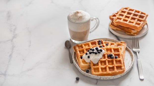 Delicious homemade baked belgian waffles with greek yogurt, blueberries and cappuccino on white marble background. Perfect breakfast with copy space for text or design