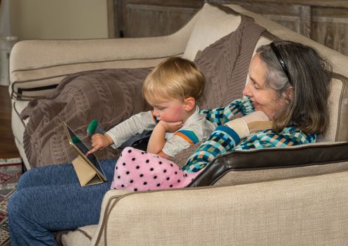 Grandmother helping small baby boy with app on a digital tablet while sitting in family room and relaxing