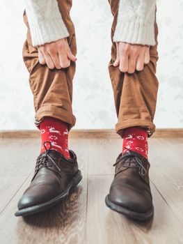 Young man pulls up leg of his chinos trousers to show bright red socks with reindeers on them. Scandinavian pattern. Winter holiday spirit. Casual outfit for New Year and Christmas celebration.