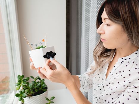 Smiling woman shows handmade decorations for Halloween. DIY flags and Boo! sticker on flowerpot with succulent plant. Socially-Distanced Halloween at home.