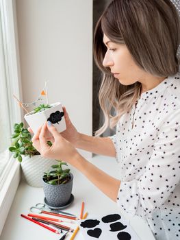 Smiling woman shows handmade decorations for Halloween. DIY flags and Boo! sticker on flowerpot with succulent plant. Socially-Distanced Halloween at home.