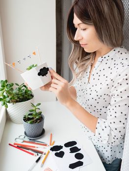 Smiling woman shows handmade decorations for Halloween. DIY flags and Boo! sticker on flowerpot with succulent plant. Socially-Distanced Halloween at home.