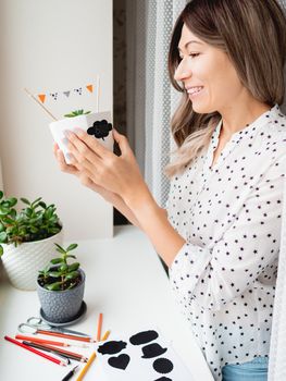 Smiling woman shows handmade decorations for Halloween. DIY flags and Boo! sticker on flowerpot with succulent plant. Socially-Distanced Halloween at home.