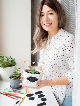 Smiling woman shows handmade decorations for Halloween. DIY flags and Boo! sticker on flowerpot with succulent plant. Socially-Distanced Halloween at home.
