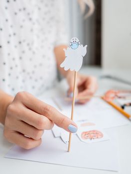 Woman with blue polish on nails paints ghost in medical protective mask. Handmade decorations for Halloween. DIY stickers on for flower pots. Socially-Distanced Halloween at home.