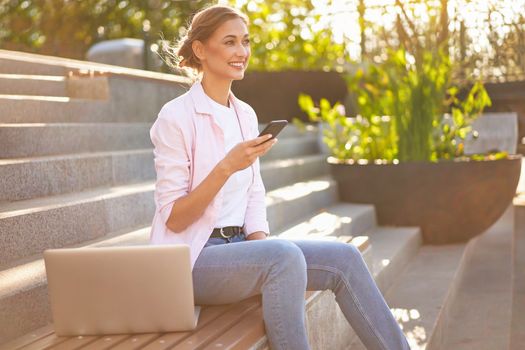 Businesswoman sitting stairs summer park using laptop and smartphone Business person working remote. Outdoor. Successful Young European caucasian woman freelancer meeting online