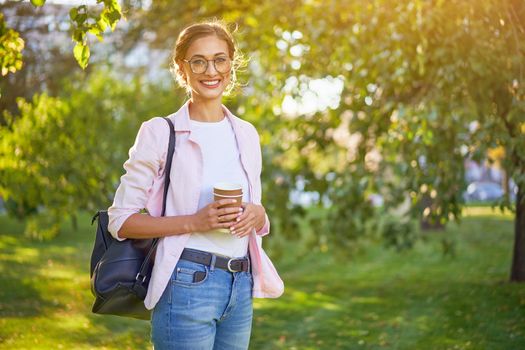 Businesswoman standing summer park Business person Outdoors Successful european caucasian woman freelancer or teacher walking outside with black backpack dressed denim jeans white shirt pink jacket