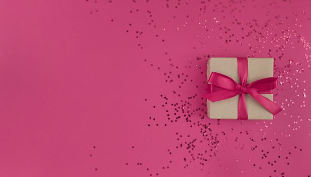 Gift box wrapped in craft paper with pink ribbon with bow and confetti on pink background. Monochrome festive flat lay with copy space.