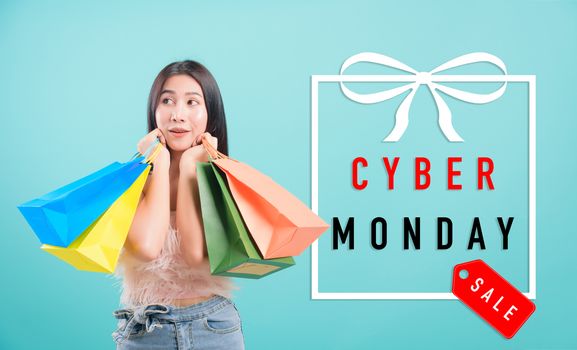 Asian happy portrait beautiful young woman standing smile in summer shopping her holding multicolor shopping bags on hand and looking to Cyber Monday text in gift box on side on blue background