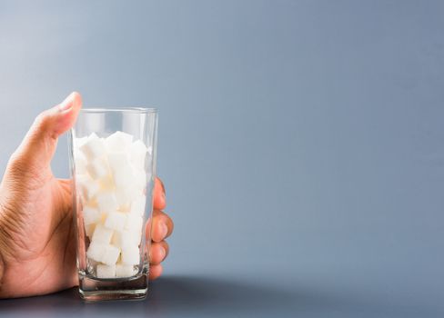 Hand hold on glass full of white sugar cube sweet food ingredient, isolated on gray background, health high blood risk of diabetes and calorie intake concept and unhealthy drink
