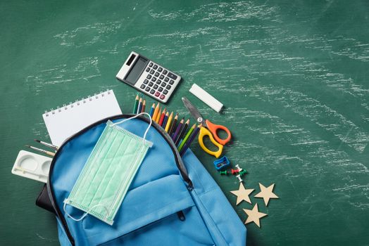 Top view of stylish blue school bag backpack at a green chalkboard with face mask protection and stationery, Back to school education new normal during outbreak COVID-19 or coronavirus concept