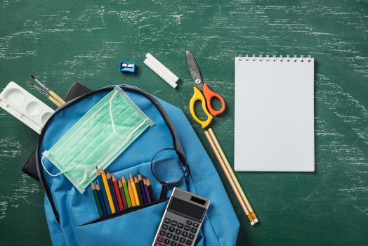 Top view of blue school bag backpack at a green chalkboard with face mask protection and have copy space for your text, Back to school education during epidemic coronavirus concept