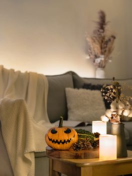 Cozy scandinavian style halloween interior. Bright orange pumpkin with Jack-o-lantern laughing face, candles on sofa background. Vertical. Copy space.