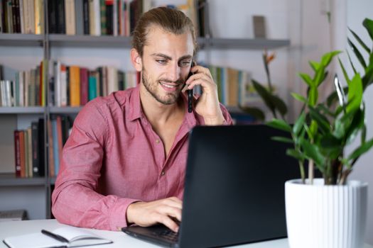 Handsome young caucasian man using computer working at home and talking with mobile feeling happy with smile showing teeth